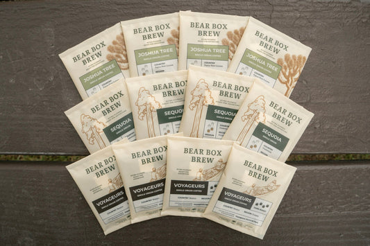 A fan arrangement of ten Bear Box Brew coffee bags on a dark weathered wooden surface. The packaging is cream-colored with green and dark brown accents, featuring the 'Bear Box Brew' single origin sachets.