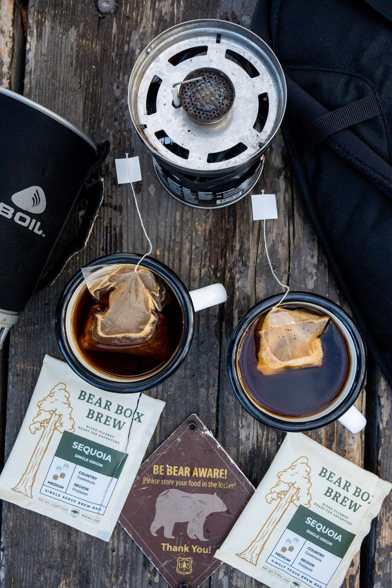 Two cups of Bear Box Brew coffee, a camping stove, and bear safety cards on a wooden surface.