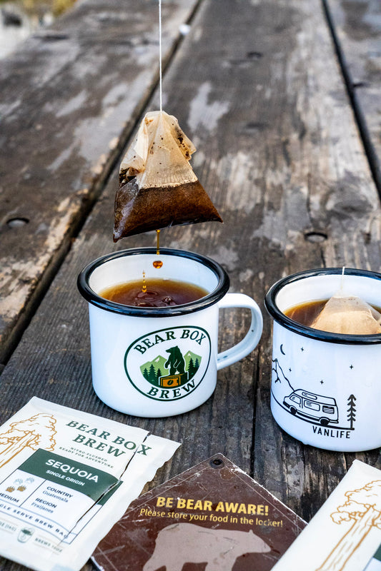 Two mugs of Bear Box Brew coffee on a wooden surface with coffee bags and packaging.