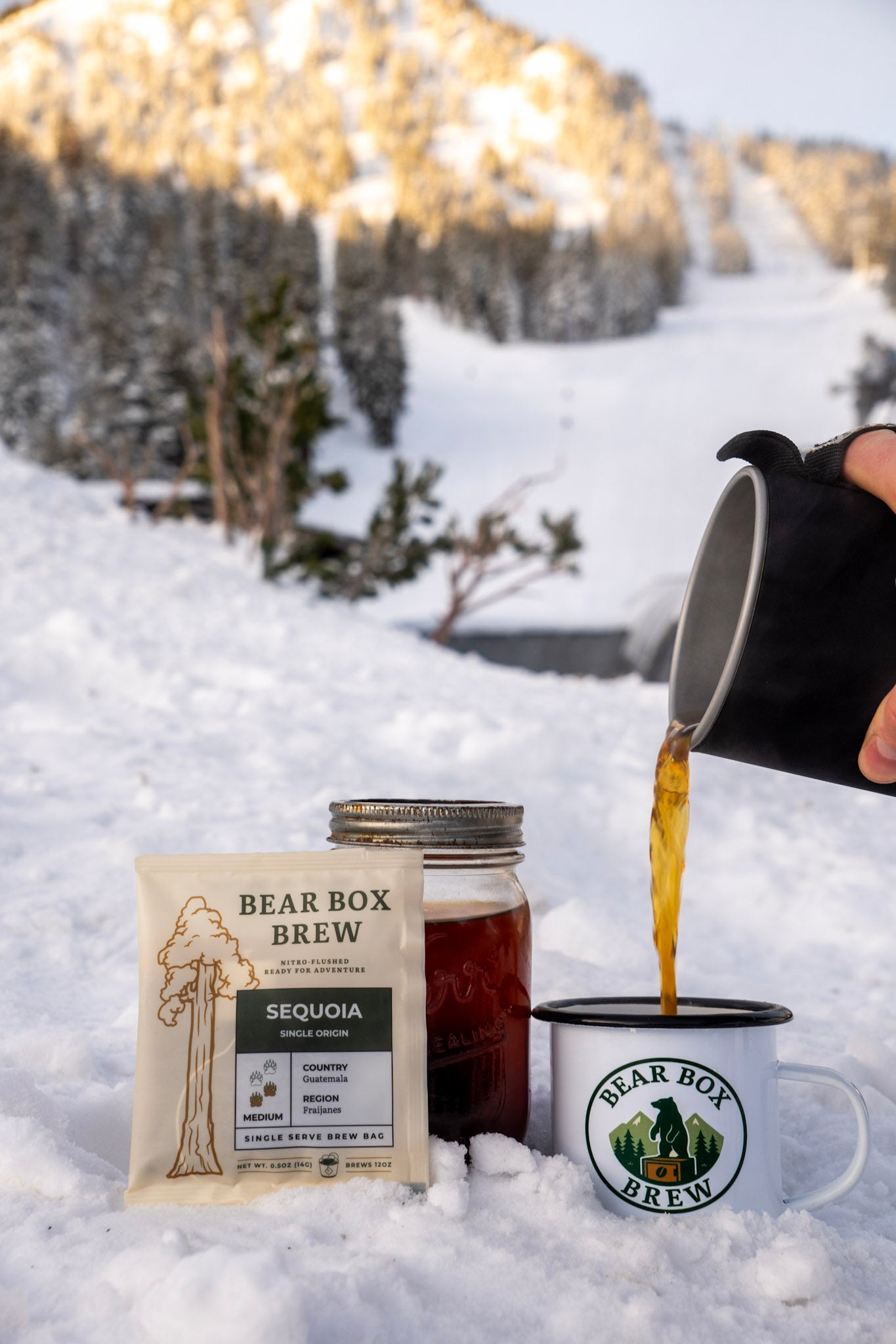 Person pouring coffee from a pot into a mug with Bear Box Brew packaging in a snowy landscape.